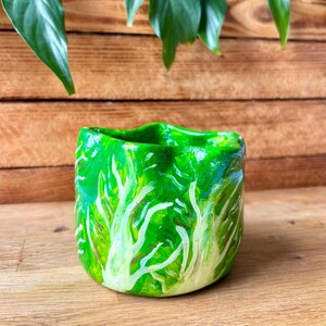May include: A small, hand-painted ceramic planter with a vibrant green and yellow lettuce leaf design. The pot has a wavy rim and is set against a wooden background, ideal for small plants or succulents.