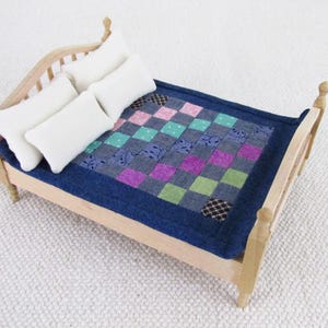 May include: A miniature wooden bed with a blue quilt featuring a patchwork design in pink, green, and purple squares. Three white rectangular pillows rest on the bed. The bed frame is light-coloured wood.