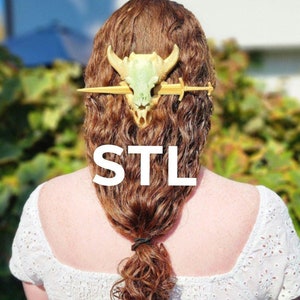 May include: A gold and green hair accessory with a skull and sword design. The word "STL" is displayed in white block letters. The accessory is worn in the hair.