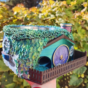 May include: A miniature hobbit house planter with a round purple door, green roof, and ivy detailing. The planter has a small chimney and a brown picket fence. The design is detailed and colourful, with a whimsical aesthetic.