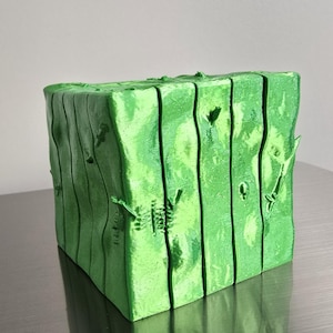 May include: A bright green, cube-shaped decorative object with a textured, wood-like appearance. The surface features vertical grooves and small, irregular cutouts. The object is displayed on a reflective, metallic surface.