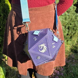 May include: A purple, multi-sided dice-shaped crossbody bag with silver hardware and a light blue strap. The bag features the numbers 2, 8, and 14, and a silver emblem. The bag is worn over the shoulder.