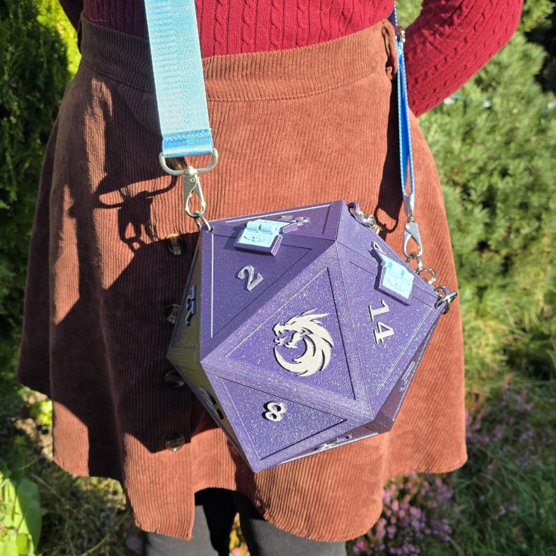 May include: A purple, multi-sided dice-shaped crossbody bag with silver hardware and a light blue strap. The bag features the numbers 2, 8, and 14, and a silver emblem. The bag is worn over the shoulder.