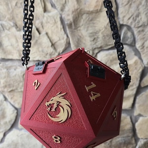May include: A red, multi-sided dice-shaped bag with a black chain strap. The bag features gold-colored numbers and a gold dragon emblem. The bag has a textured surface and a clasp closure. The numbers on the bag are visible as 2, 8, 14, and 19.