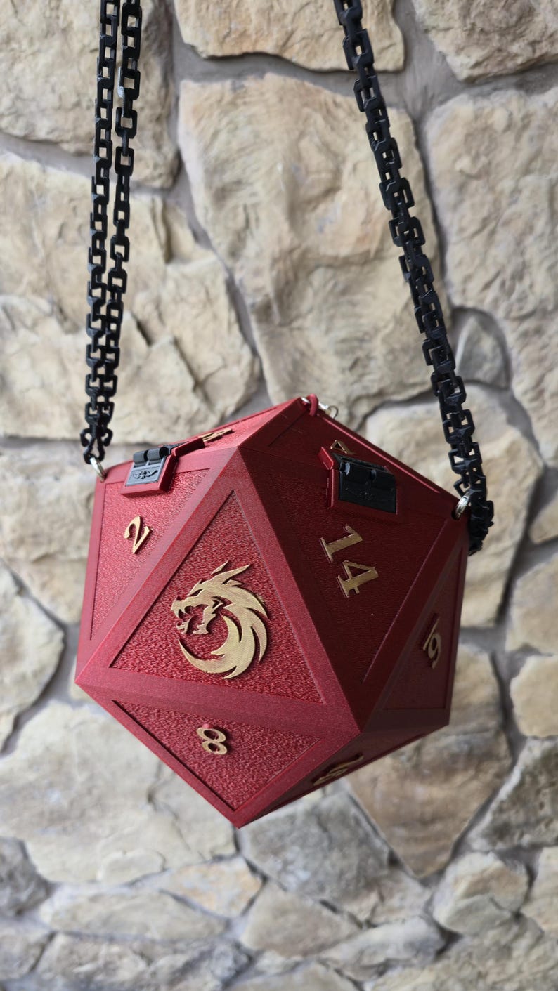 May include: A red, multi-sided dice-shaped bag with a black chain strap. The bag features gold-colored numbers and a gold dragon emblem. The bag has a textured surface and a clasp closure. The numbers on the bag are visible as 2, 8, 14, and 19.