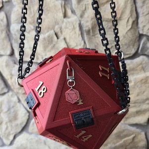 May include: A red, geometric dice-shaped box with the numbers 11, 14, and 18 visible. The box is suspended by black chain links. A small charm with a key and a geometric shape hangs from the box. The box has a hinged lid.