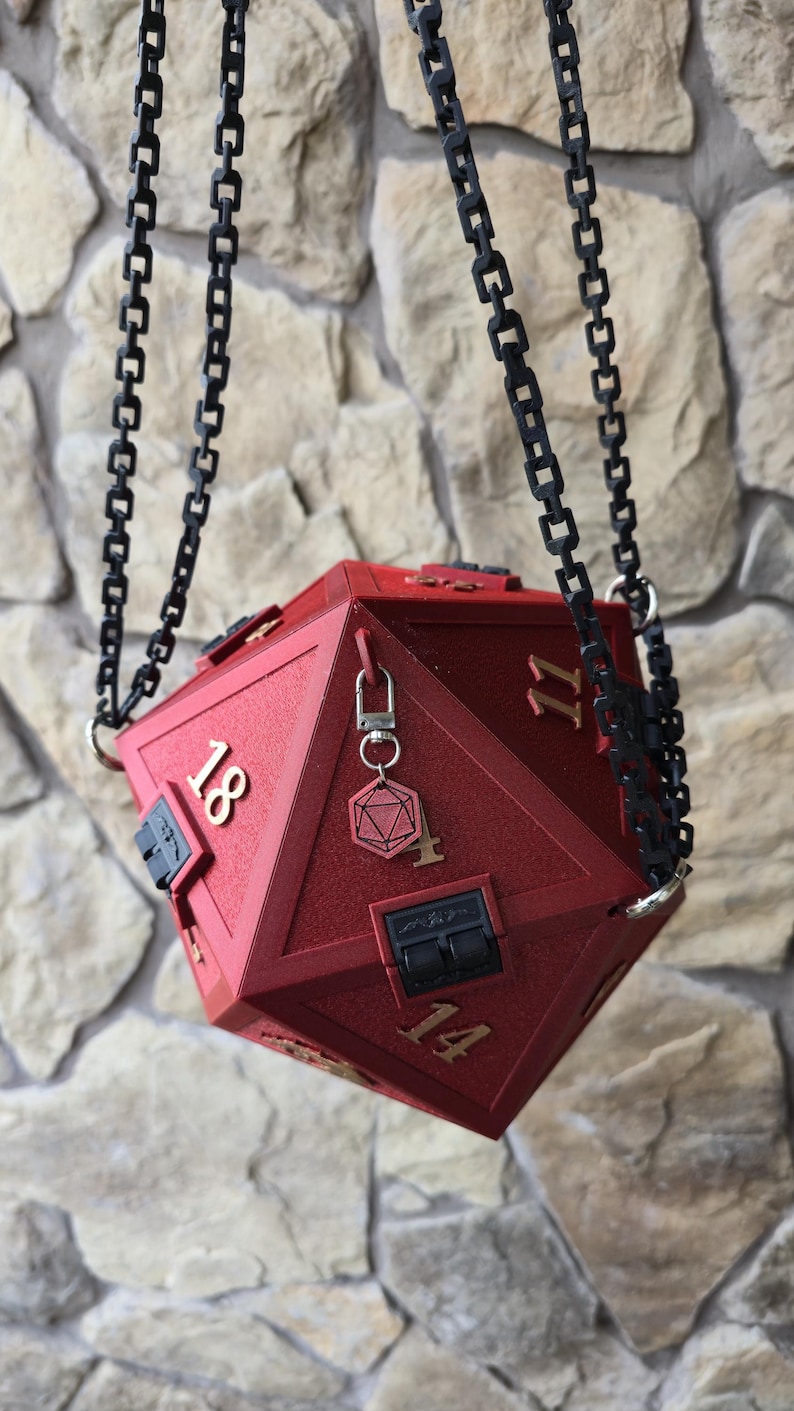 May include: A red, geometric dice-shaped box with the numbers 11, 14, and 18 visible. The box is suspended by black chain links. A small charm with a key and a geometric shape hangs from the box. The box has a hinged lid.