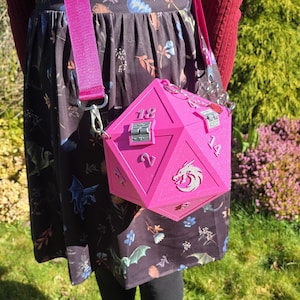May include: A bright pink, geometric-shaped handbag with silver hardware and a matching pink strap. The bag is designed to resemble a twenty-sided die, with numbers and a dragon emblem on its faces. The bag is worn over the shoulder.