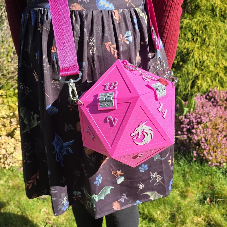 May include: A bright pink, geometric-shaped handbag with silver hardware and a matching pink strap. The bag is designed to resemble a twenty-sided die, with numbers and a dragon emblem on its faces. The bag is worn over the shoulder.