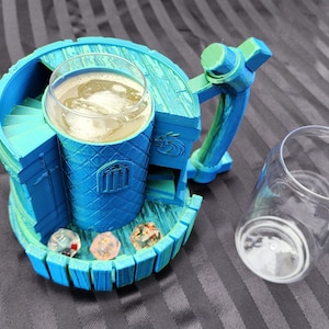 May include: A blue and green 3D printed tower-shaped mug with a handle shaped like a drawbridge. The mug has a clear glass insert and is filled with a light brown beverage with ice. There are four multi-coloured dice on the base of the mug.