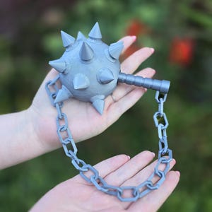 May include: A grey, spiked mace with a chain, held in open hands against a blurred green and red background. The mace head is round with conical spikes. The chain is made of interlocking links.