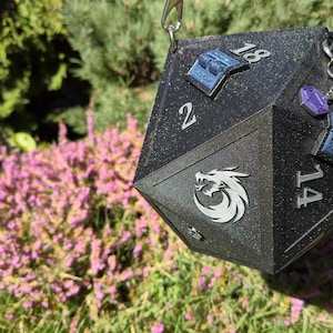 May include: A black, glittery D20 dice-shaped bag with silver chain straps. The bag features white numbers, a dragon logo, and decorative blue and purple accents. The background includes green foliage and pink flowers.