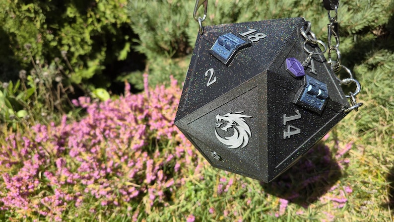 May include: A black, glittery D20 dice-shaped bag with silver chain straps. The bag features white numbers, a dragon logo, and decorative blue and purple accents. The background includes green foliage and pink flowers.