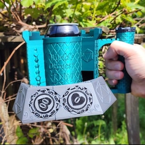 May include: A teal green 3D printed can holder shaped like a castle with a silver metal base featuring two circular dice with numbers and a heart symbol. The text "Worlds Okayest DM" is printed on the side of the can holder.