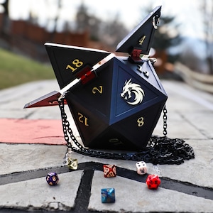 May include: Black, geometric dice-shaped handbag with gold numbering and a chain strap. The bag has an open top and a gold dragon emblem. Several multi-colored dice are scattered around the bag on a stone surface.