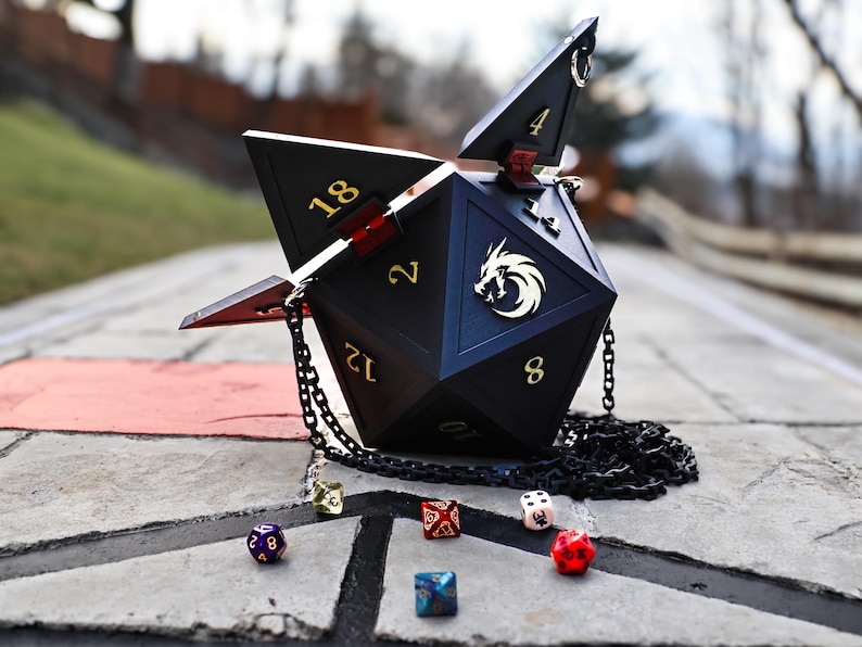 May include: Black, geometric dice-shaped handbag with gold numbering and a chain strap. The bag has an open top and a gold dragon emblem. Several multi-colored dice are scattered around the bag on a stone surface.