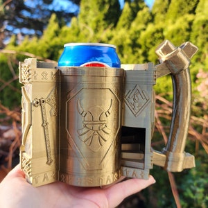 Könnte beinhalten: Ein goldfarbener 3D-gedruckter Dosenhalter in Form eines Wikingerbechers mit Henkel. Der Dosenhalter hat ein Wikingerhelm-Design und Runen an der Seite. Eine blaue Dose befindet sich im Halter.