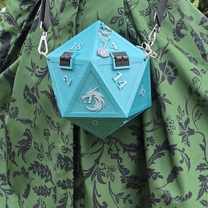 May include: A light blue, geometric-shaped handbag with the numbers 2, 4, 8, 14, and 18 visible. The bag has a silver chain strap and a silver charm. The bag has a silver emblem of a griffin. The bag is hanging in front of a green dress with a black floral pattern.