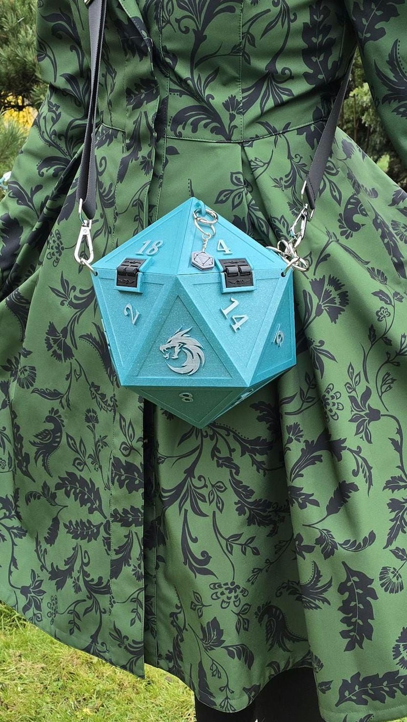 May include: A light blue, geometric-shaped handbag with the numbers 2, 4, 8, 14, and 18 visible. The bag has a silver chain strap and a silver charm. The bag has a silver emblem of a griffin. The bag is hanging in front of a green dress with a black floral pattern.