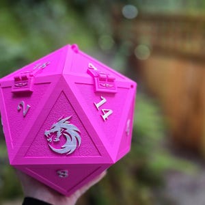 May include: A bright pink, multi-sided die with silver numerals and a silver emblem. The die has a hinged lid and is designed to hold items. The background is blurred, with green foliage and a wooden fence visible.
