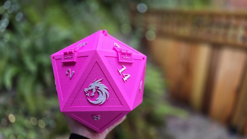 May include: A bright pink, multi-sided die with silver numerals and a silver emblem. The die has a hinged lid and is designed to hold items. The background is blurred, with green foliage and a wooden fence visible.
