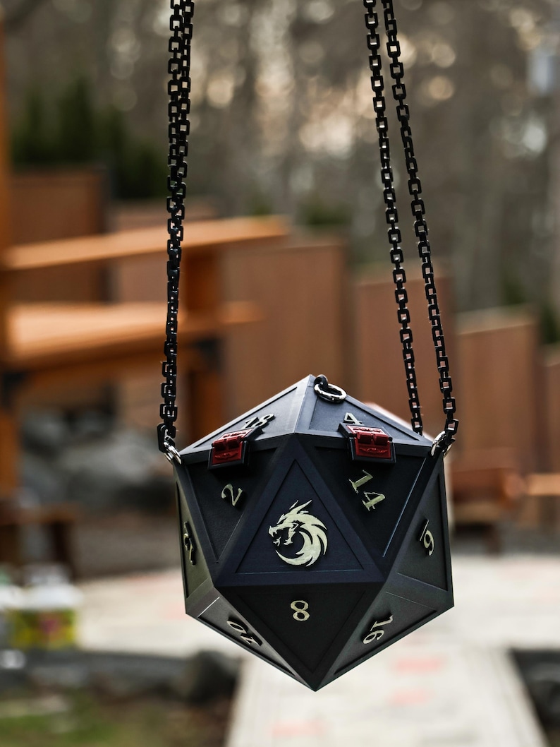 May include: A black, multi-sided dice-shaped bag with a chain handle. The bag features gold-colored numbers and a dragon emblem. Red accents are visible on the top. The bag is designed for role-playing games.