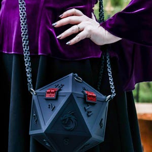 May include: A dark gray, geometric-shaped handbag with red accents and a silver chain strap. The bag features a dragon emblem and numerical markings, resembling a twenty-sided die. The bag is held by a person wearing a purple velvet top.