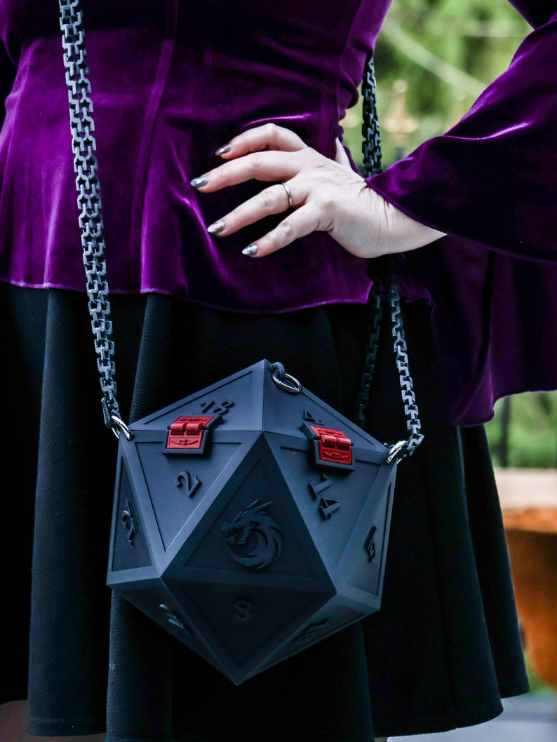 May include: A dark gray, geometric-shaped handbag with red accents and a silver chain strap. The bag features a dragon emblem and numerical markings, resembling a twenty-sided die. The bag is held by a person wearing a purple velvet top.
