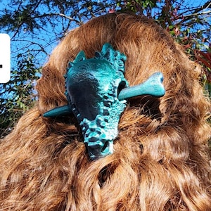 May include: A teal and black skull hair clip with a bone-shaped pin. The clip is worn in a person's hair.