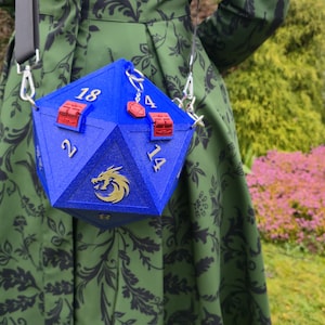 May include: A blue, multi-sided bag with silver accents, resembling a large die. The bag has red clasps and silver numbers. A gold dragon emblem is on the front. The bag is suspended from a black strap.
