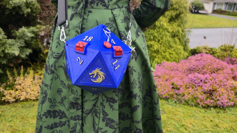 May include: A blue, multi-sided bag with silver accents, resembling a large die. The bag has red clasps and silver numbers. A gold dragon emblem is on the front. The bag is suspended from a black strap.