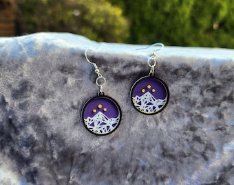 Könnte beinhalten: Ein Paar violette und weiße Ohrringe mit einem Berg- und Sternendesign. Die Ohrringe sind aus Holz und haben silberne Haken.