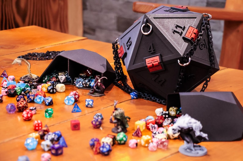 May include: A black, geometric dice bag and dice set, with a large D20 bag and a smaller dice tray. The D20 bag has red accents and a chain strap. Assorted colorful dice and miniature figurines are scattered around the bags on a wooden surface.
