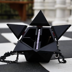 May include: Black geometric handbag with a chain strap, open to reveal a smartphone and pen. The bag has a unique, angular design with multiple triangular panels. The background is a black and white checkered pattern.