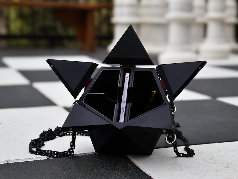 May include: Black geometric handbag with a chain strap, open to reveal a smartphone and pen. The bag has a unique, angular design with multiple triangular panels. The background is a black and white checkered pattern.
