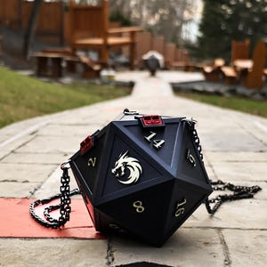 May include: Black, multi-sided dice-shaped handbag with gold numbers and a white dragon emblem. The bag has a chain strap and red accents. A unique accessory for fans of tabletop games.