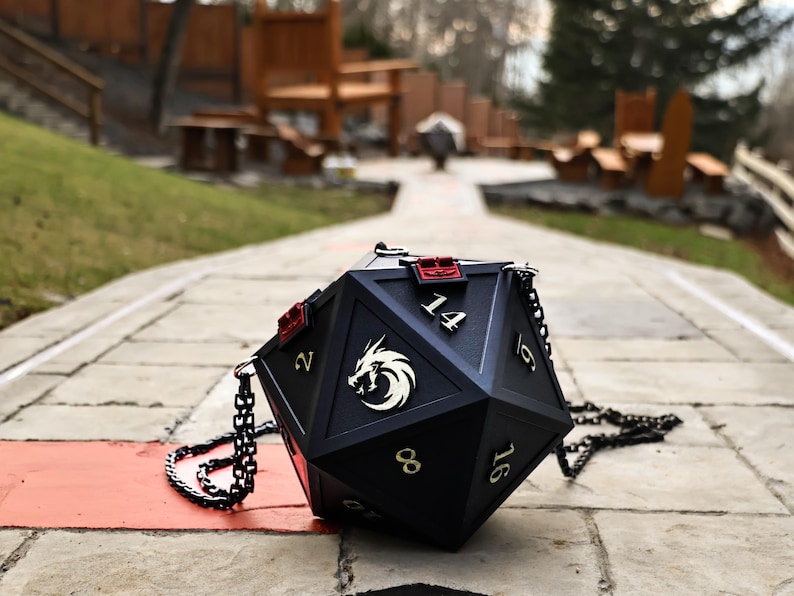 May include: Black, multi-sided dice-shaped handbag with gold numbers and a white dragon emblem. The bag has a chain strap and red accents. A unique accessory for fans of tabletop games.