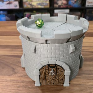 Puede incluir: Una torre de dados gris impresa en 3D con forma de torreón de castillo. La torre tiene una puerta de madera y una parte superior almenada. Un dado verde y amarillo de múltiples caras descansa en la parte superior. La torre está diseñada para juegos de mesa.