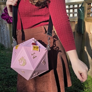 May include: A pink, geometric D20 dice-shaped handbag with gold accents and a dragon emblem. The bag has a black shoulder strap and silver hardware. The numbers 2, 14, 16, and 18 are visible on the dice. The bag is worn over a brown corduroy skirt.