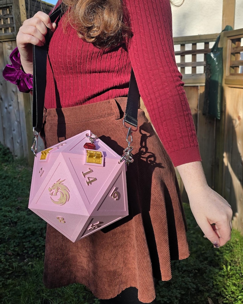 May include: A pink, geometric D20 dice-shaped handbag with gold accents and a dragon emblem. The bag has a black shoulder strap and silver hardware. The numbers 2, 14, 16, and 18 are visible on the dice. The bag is worn over a brown corduroy skirt.