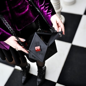 May include: A black, geometric, multi-sided handbag with a chain strap. The bag is held open, revealing the interior. The person is wearing a purple velvet jacket and black boots, standing on a black and white checkered surface.