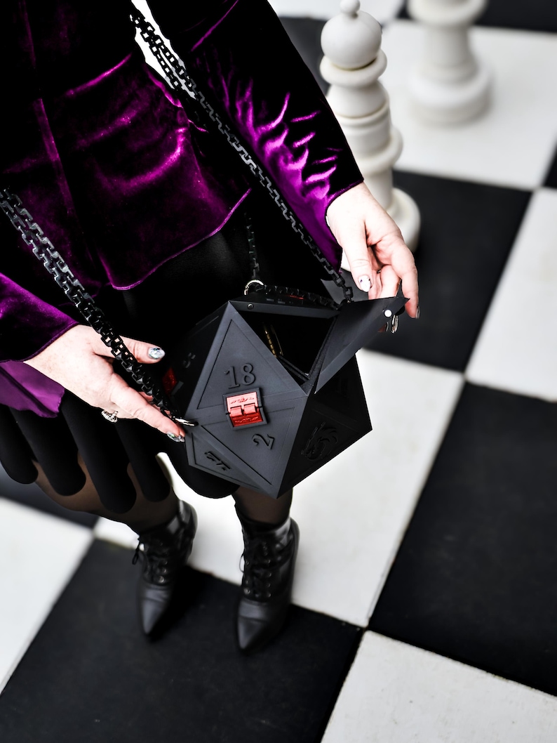 May include: A black, geometric, multi-sided handbag with a chain strap. The bag is held open, revealing the interior. The person is wearing a purple velvet jacket and black boots, standing on a black and white checkered surface.