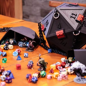 May include: A large black D20 dice bag with a chain strap and red accents, next to a smaller black dice container. Assorted colorful dice and miniature figurines are scattered on a wooden surface, suggesting a tabletop gaming theme.