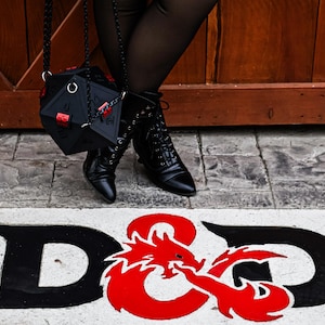 May include: Black, geometric dice-shaped handbag with red accents, hanging from a chain. The image also shows black boots and the D&D logo in black and red on a white background, suggesting a role-playing game theme.