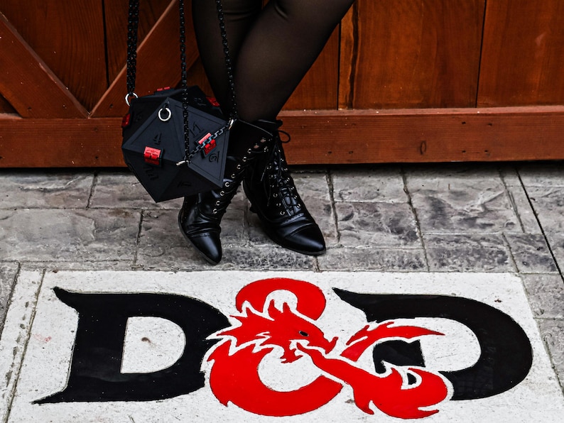 May include: Black, geometric dice-shaped handbag with red accents, hanging from a chain. The image also shows black boots and the D&D logo in black and red on a white background, suggesting a role-playing game theme.