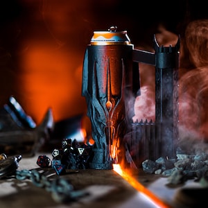 May include: A black metal can holder shaped like a demon with a can of beverage inside. The holder is surrounded by black dice and small rocks. There is smoke and fire coming from the holder.