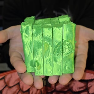 May include: A bright green, textured, rectangular object with a design on the side is held in open hands. The object appears to be made of stacked rectangular blocks. The background is blurred.