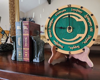 May include: A green and gold clock with a wood grain design and black hands. The clock face has the words "Second Breakfast", "Elevenses", "Lunch", "Afternoon", "Dinner", and "Supper" written around the edge. The clock is on a wooden stand.