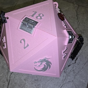 May include: A large, pink, multi-sided box resembling a die, with the numbers 2, 14, and 18 visible. It has a silver clasp and a dragon emblem. Black straps and a keychain are attached. This item is likely for tabletop gaming.