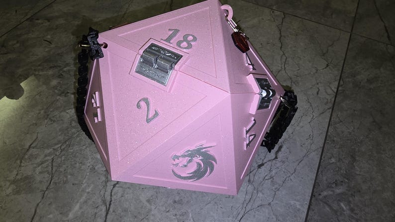 May include: A large, pink, multi-sided box resembling a die, with the numbers 2, 14, and 18 visible. It has a silver clasp and a dragon emblem. Black straps and a keychain are attached. This item is likely for tabletop gaming.